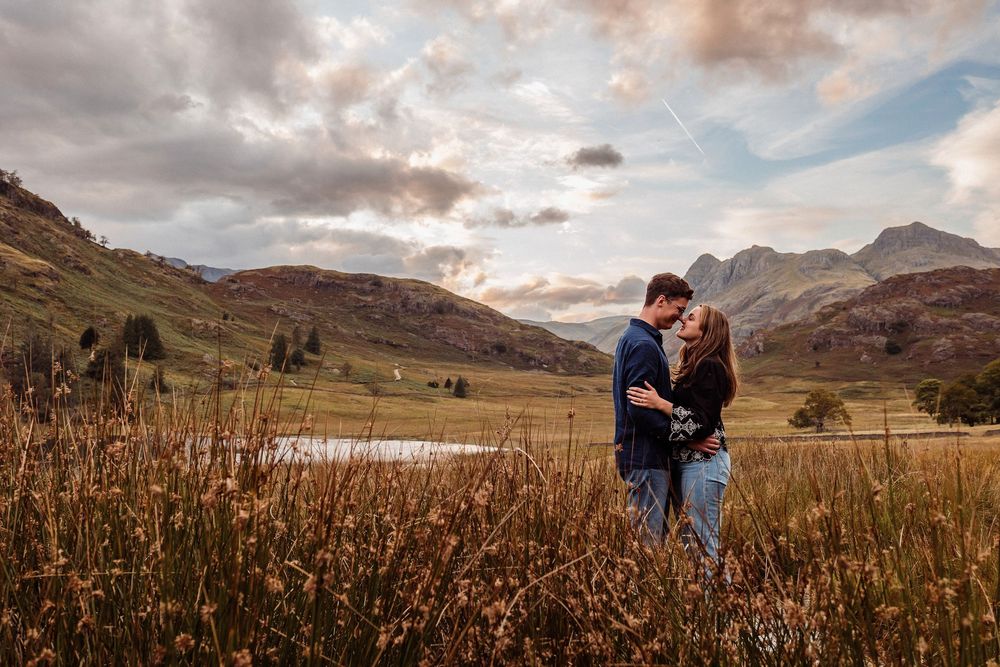 A couple embraces in a golden meadow at sunset with dramatic mountain peaks and cloudy skies in the background.