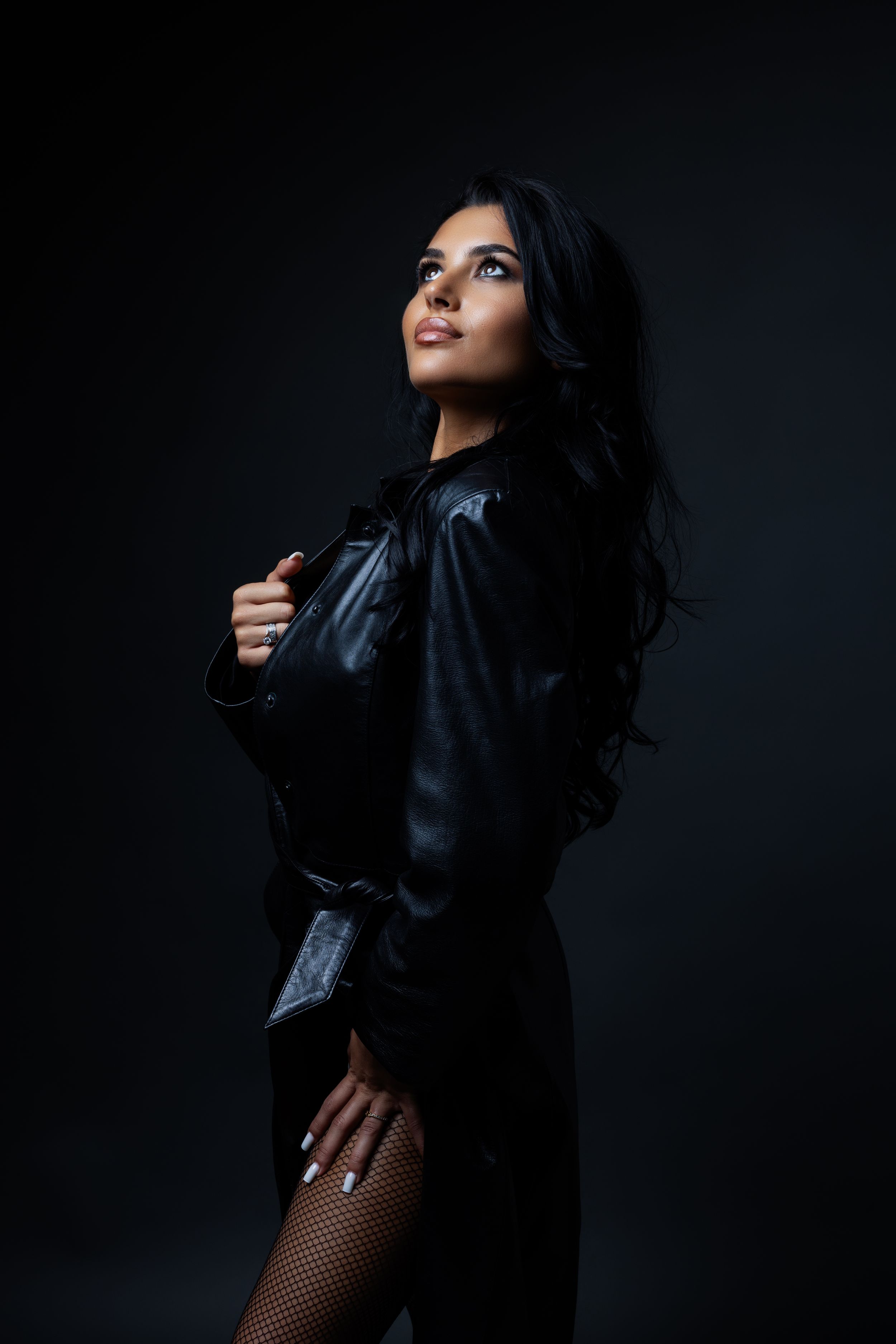 Dramatic fashion portrait series in dark lighting shows model in black jacket and fishnet stockings posing against dark backdrop.