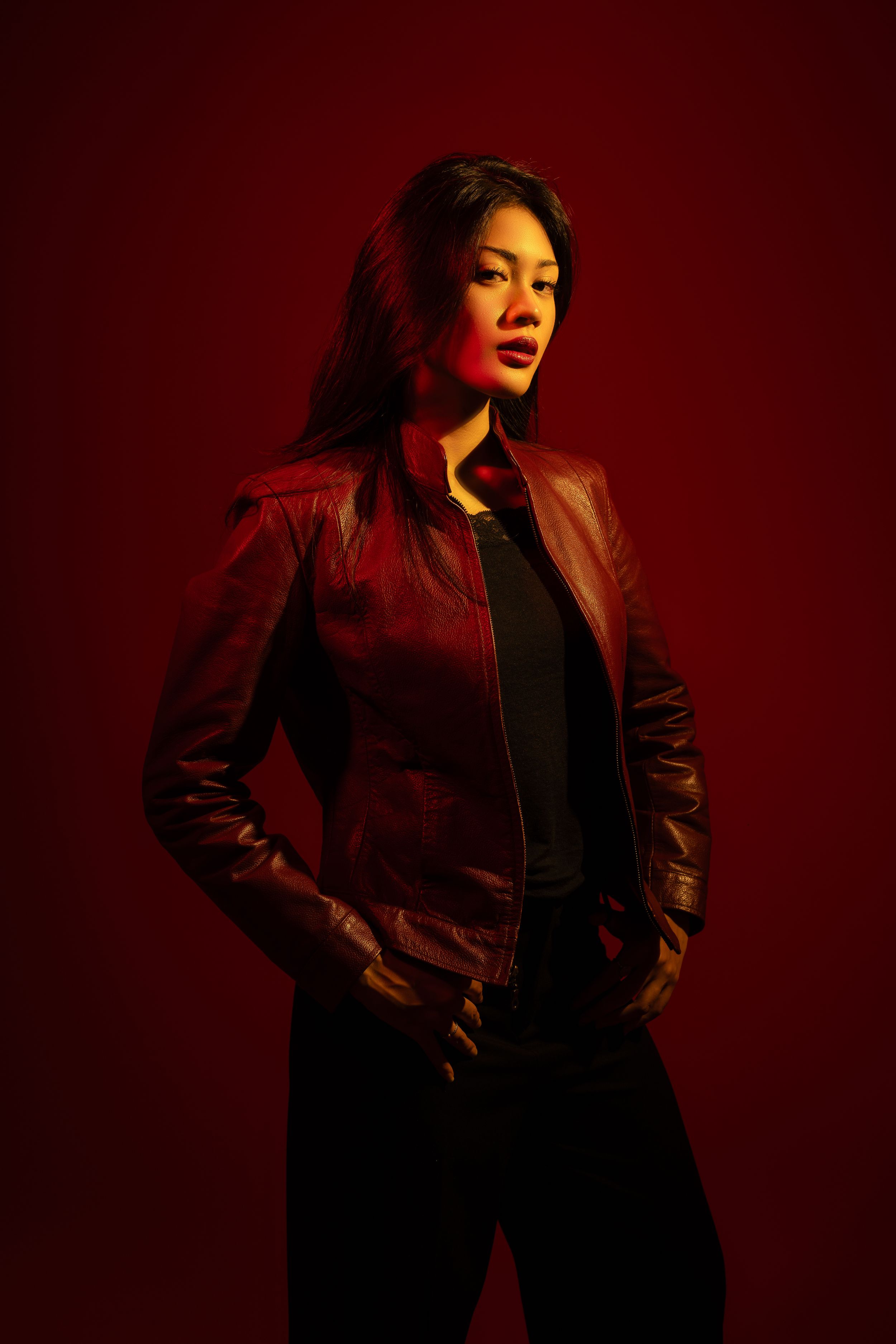 A fashion model poses in a black leather jacket against a dark red background in a dramatic photoshoot series.