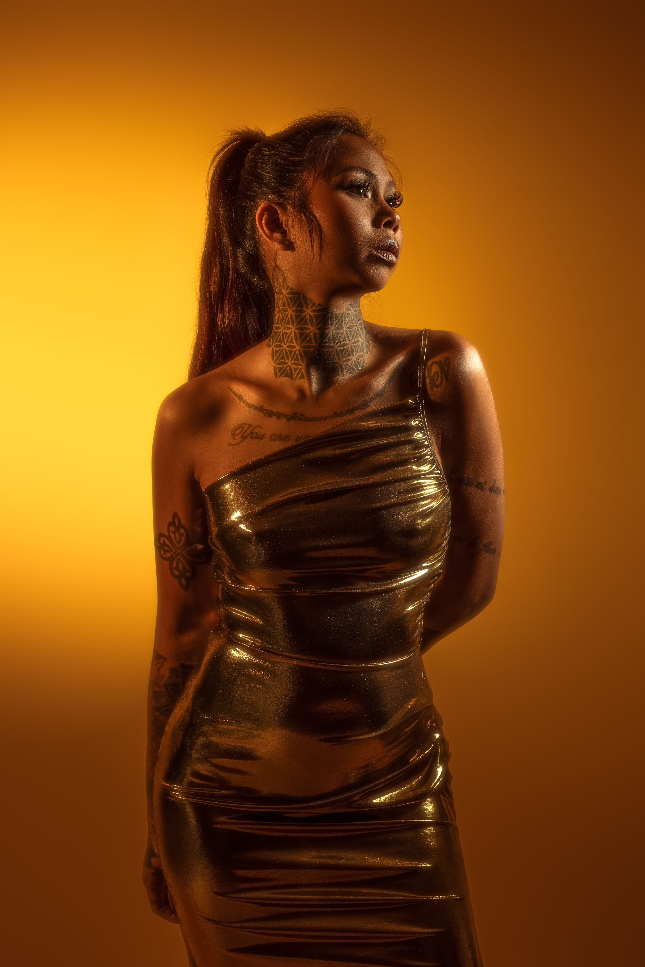 A figure in a shimmering gold dress poses dramatically against a warm amber backdrop in a fashion photoshoot series.