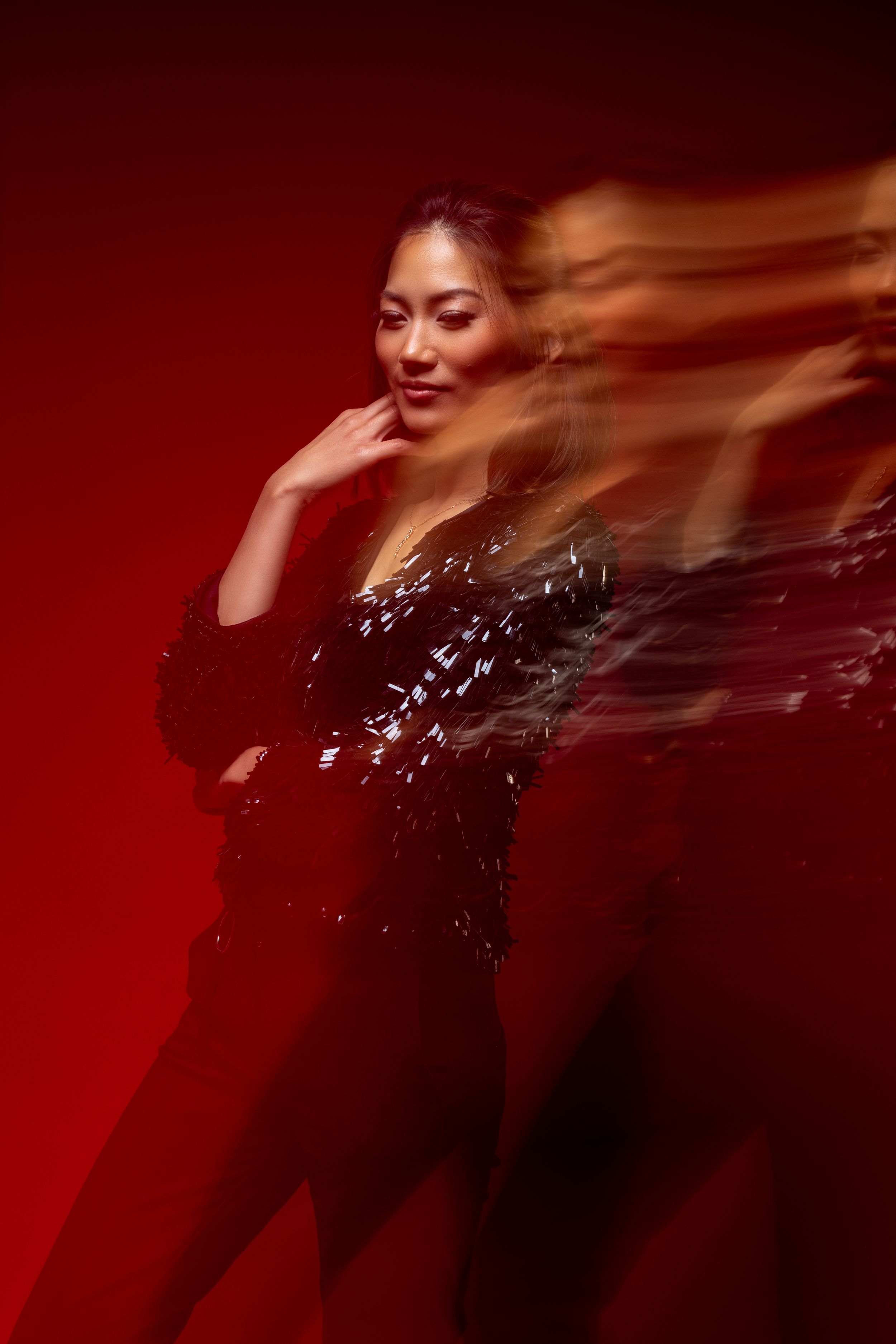 Artistic portrait with motion blur effect against deep red background showing sparkly black outfit and dramatic lighting.