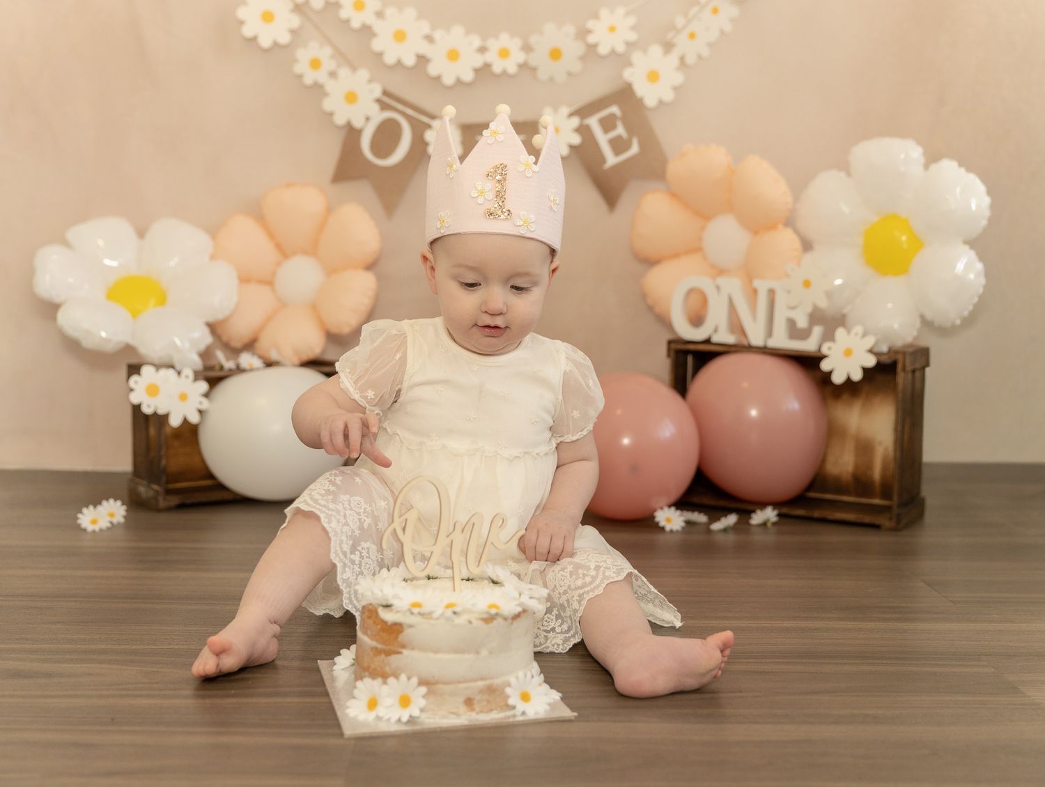 First birthday celebration setup with wicker basket, pink and brown balloons, and 'ONE' banner hanging with greenery garland.