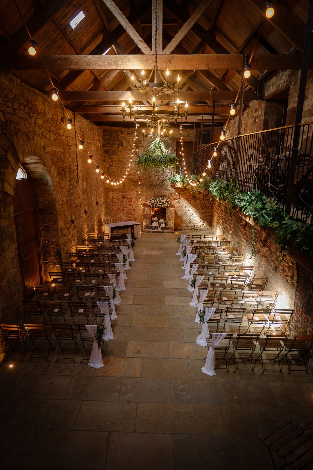 Rustic barn wedding ceremony setup with string lights and white fabric decorations on chairs.