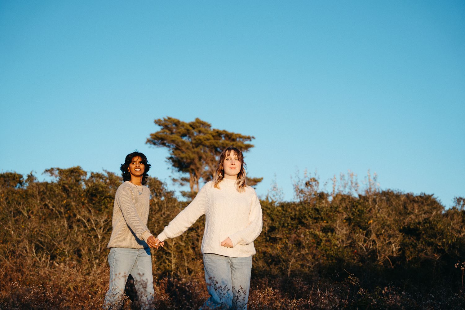 A couple stands together in a field during golden hour with trees in the background. Engagement Photos at Fort Fisher, NC