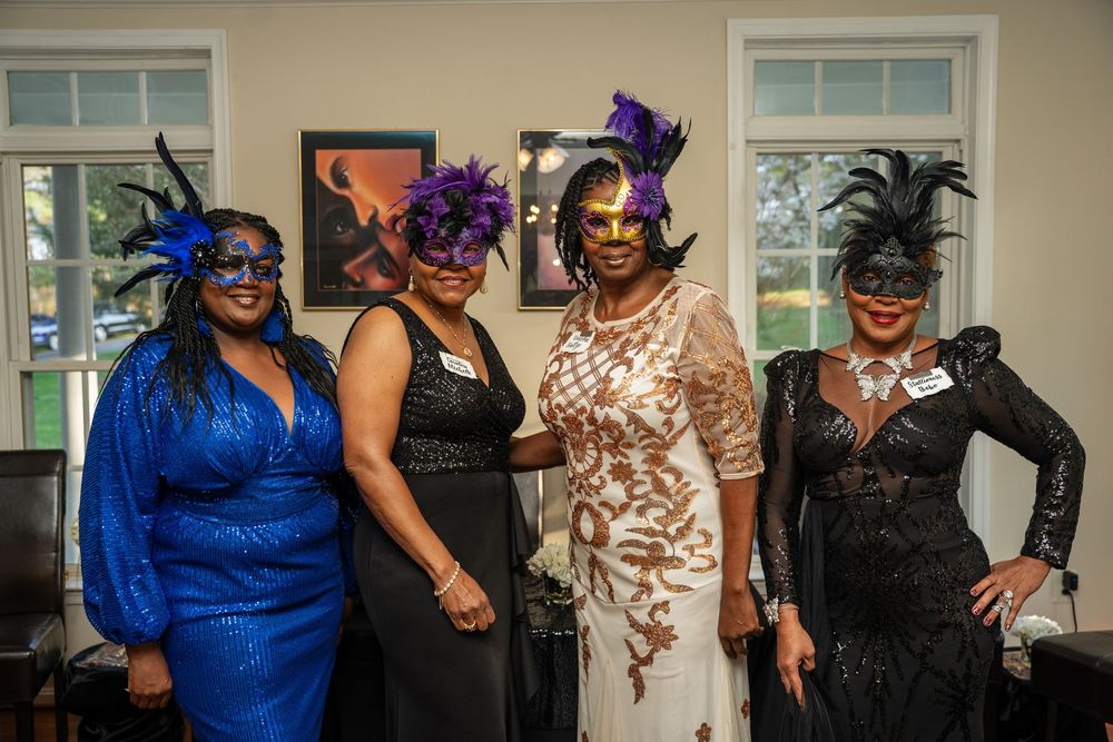 Group photo of people wearing fancy dress outfits with elaborate hats and formal attire at an indoor event.