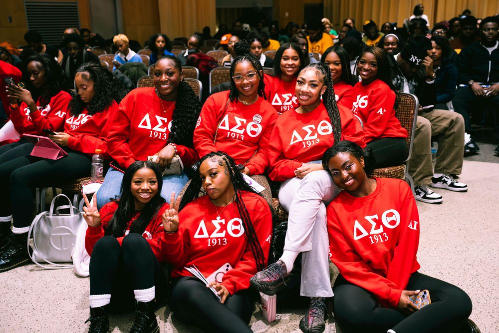 A group wearing matching bright red sweatshirts with Greek letters sits together at an indoor event.