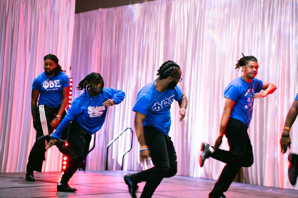 Dancers in bright blue shirts performing synchronized dance moves against pink backdrop.