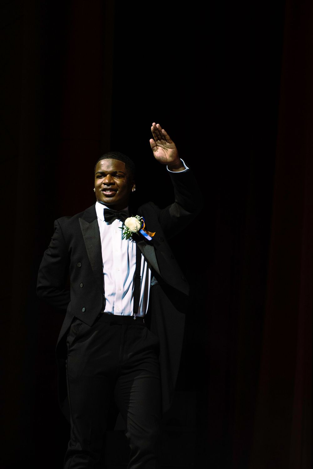A performer in a black tuxedo and white boutonniere waves to the audience while standing on a dark stage.