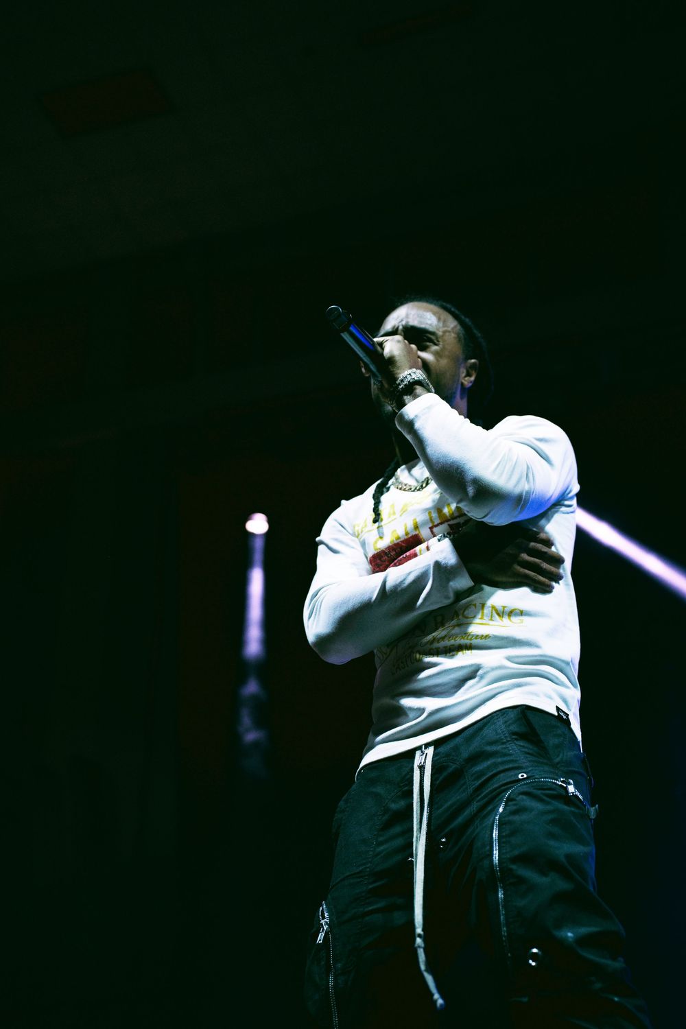 Artist performing on dimly lit stage wearing white top and dark pants with dramatic side lighting.