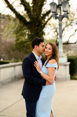 Couple embracing outdoors, man in navy suit kissing woman's cheek, she wears a light blue off-shoulder dress, trees behind.