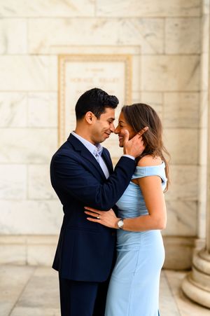 A couple in elegant attire shares a romantic moment, the man in navy suit gently touching the woman's face in a marble setting.