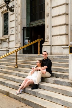 A couple sits together on the stone steps of a grand historic building with ornate architectural details.