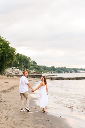 Two people walk hand in hand along a peaceful beach shoreline at sunset with trees in the background.
