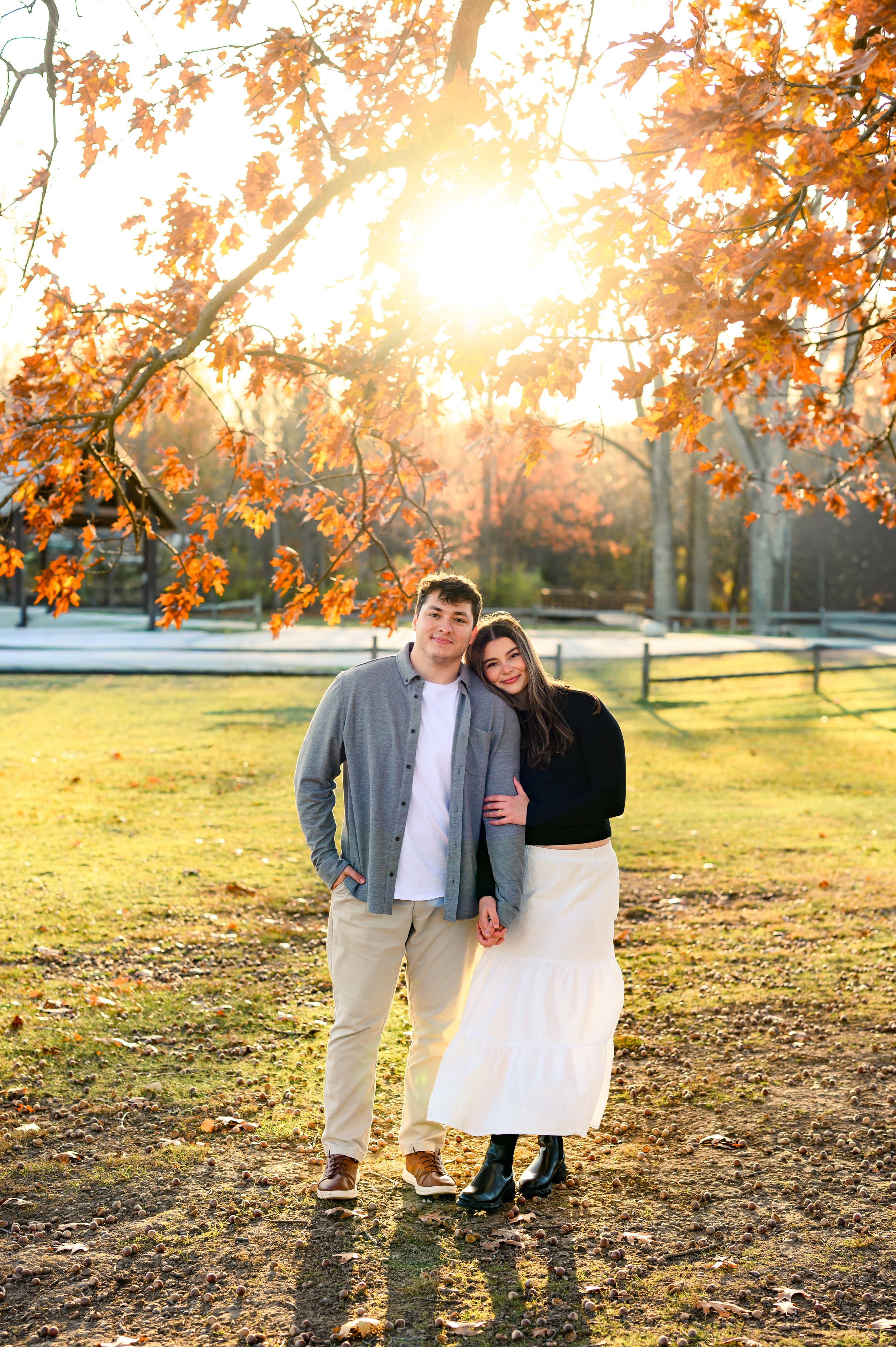 Two people stand together in a park during golden hour with autumn trees and fallen leaves in the background.