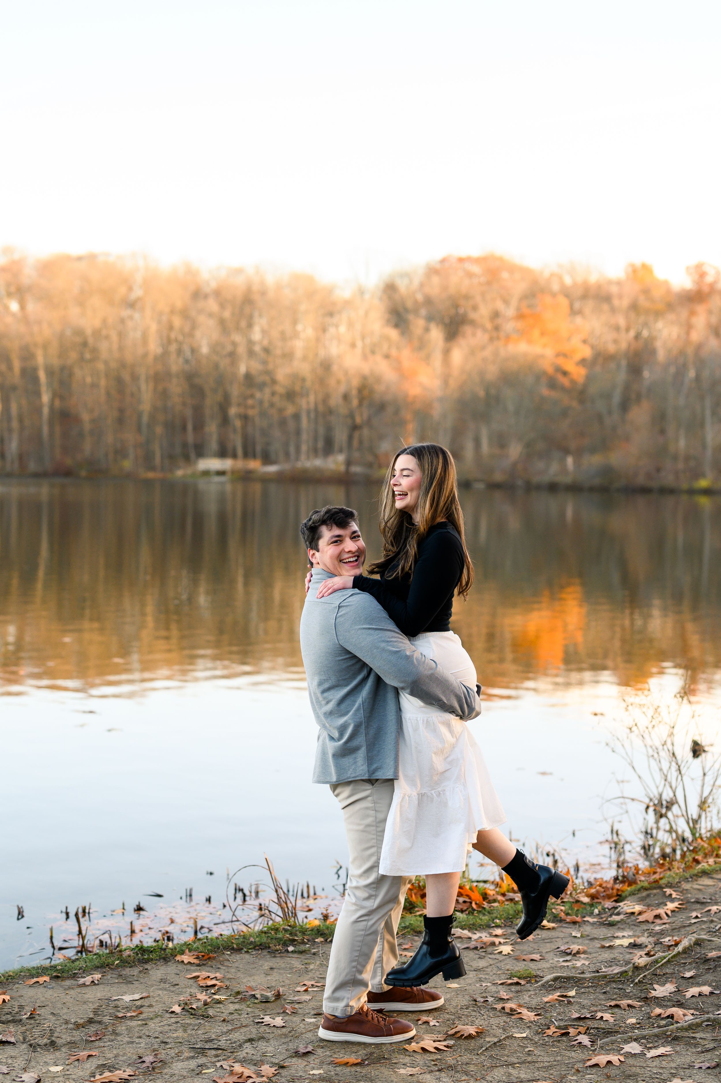 A couple embraces by a serene lake during autumn with fallen leaves scattered on the shore at sunset.