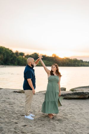 A couple dances playfully on a sandy beach at sunset with water and trees in the background.