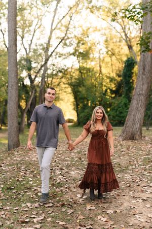 A couple walks hand in hand through a scenic autumn forest path covered in fallen leaves during golden hour.
