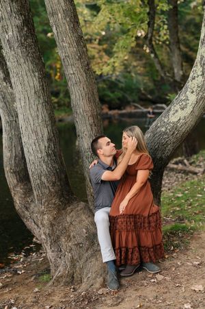 A couple shares an intimate moment together while sitting against a large tree trunk in a wooded outdoor setting.