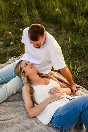 Pregnant woman lying on blanket outdoors smiling up at man in white shirt during golden hour maternity photo session.