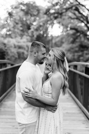 Black and white photo of a couple sharing a romantic moment on a wooden bridge surrounded by trees.