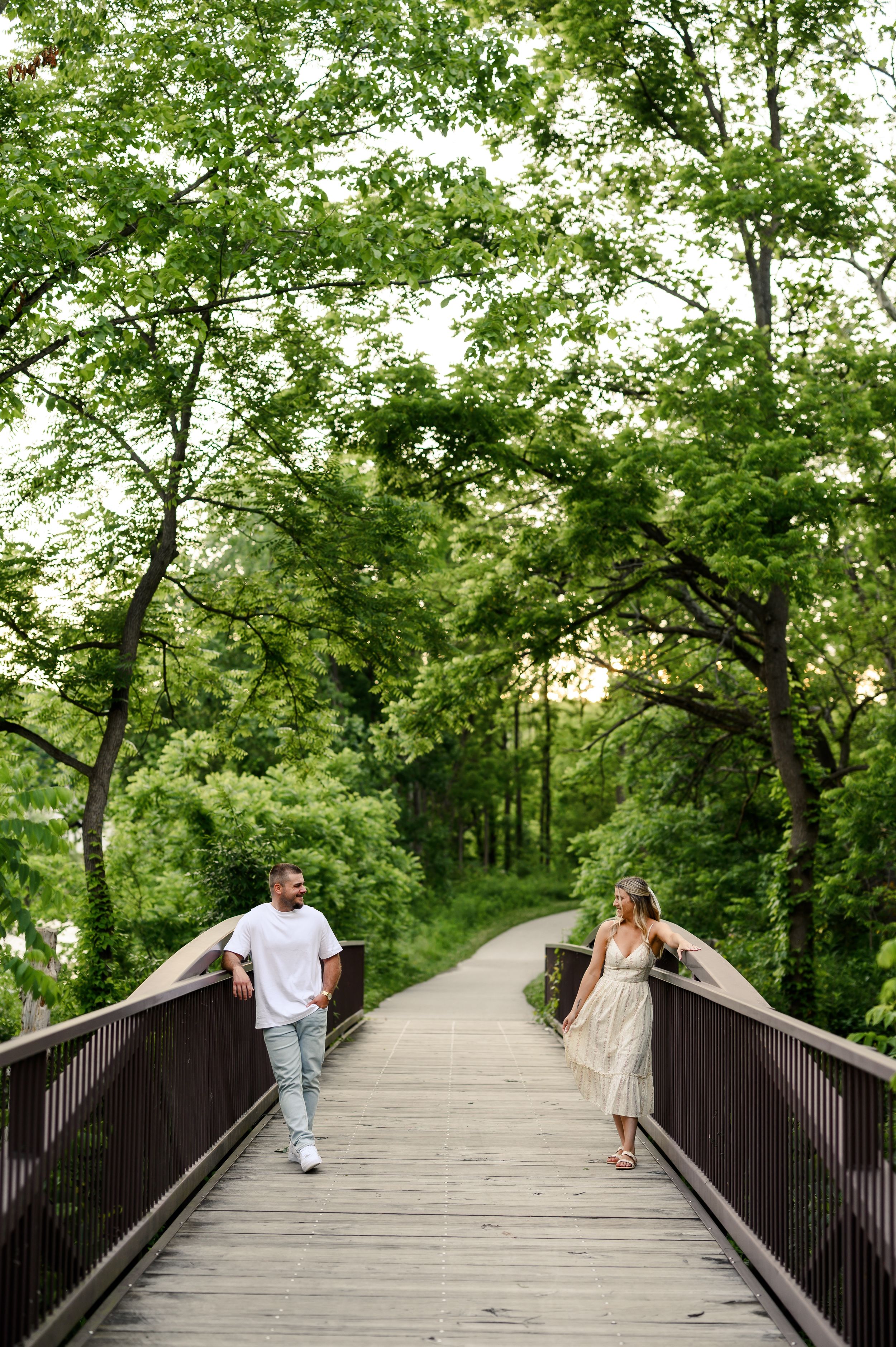 Couple walking toward each other on a wooden bridge surrounded by lush green trees in a park setting.
