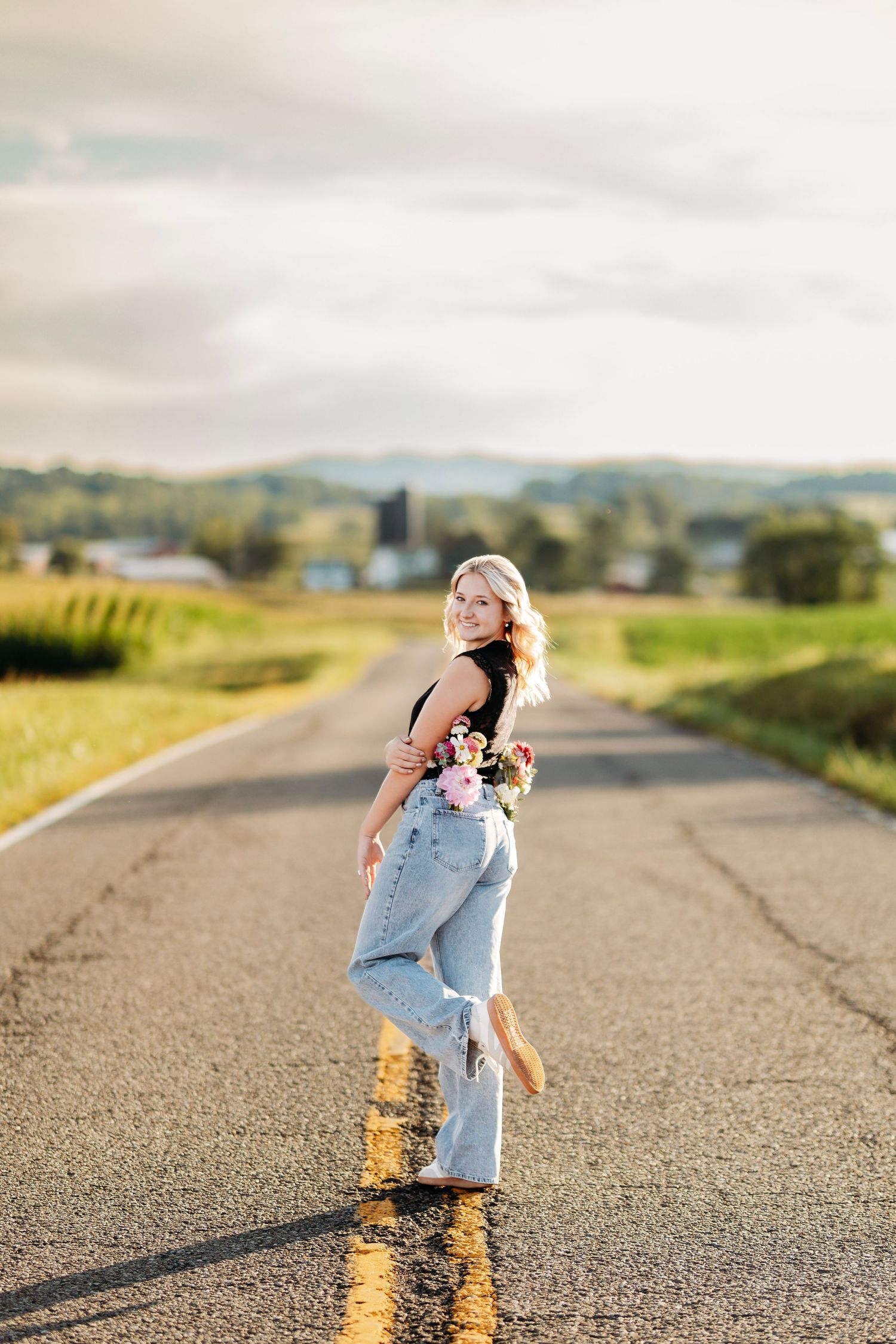 Blonde woman in jeans holding flowers posing on rural road with rolling hills at sunset.