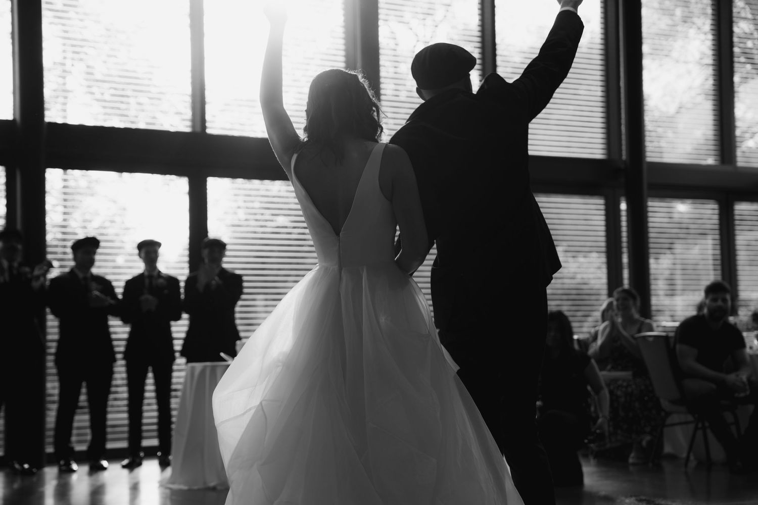 Wedding guests dance and celebrate in silhouette during a reception in a large windowed venue.