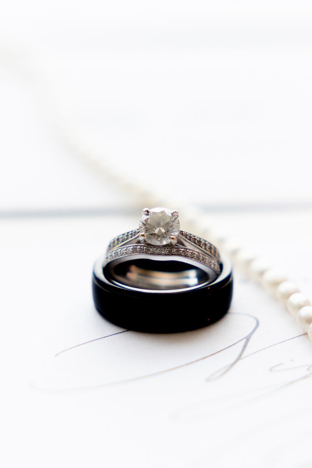 Wedding rings arranged with a diamond engagement ring and black band on a white surface with soft lighting.