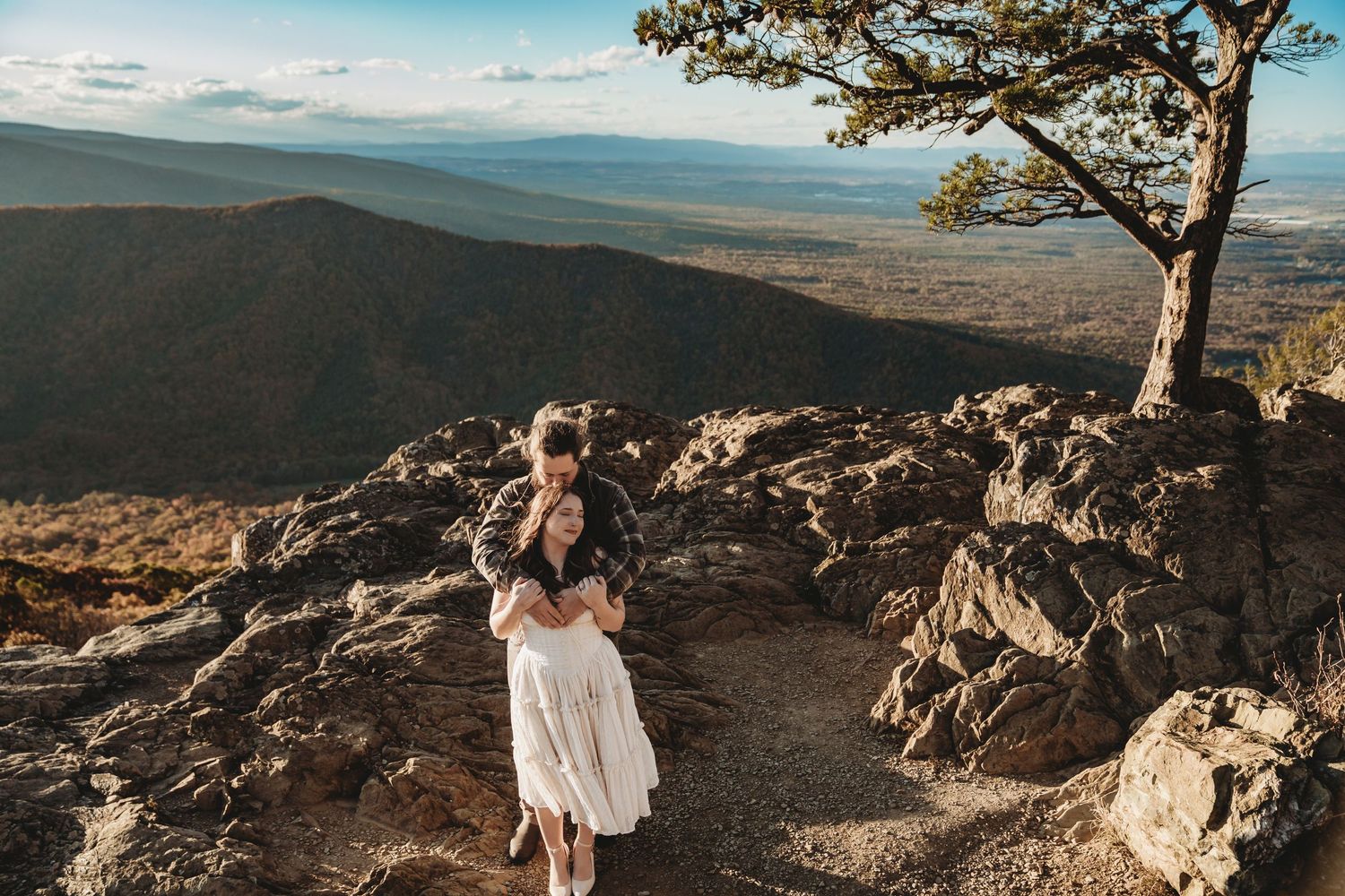 Couple embracing on rocky mountain overlook with sweeping valley views and lone tree at golden hour.