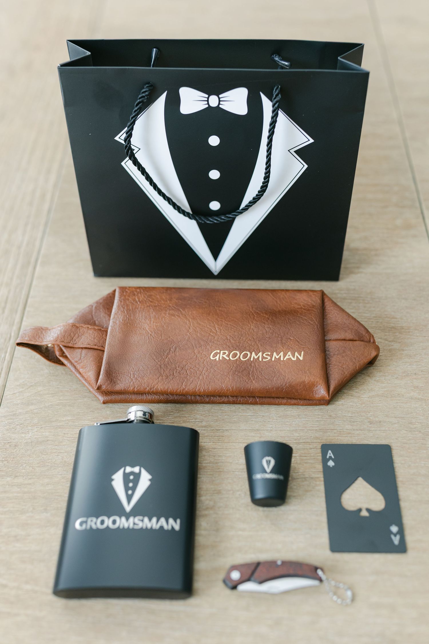 Black and brown groomsman gift set featuring tuxedo-themed bag, leather pouch, flask, playing cards, and accessories.