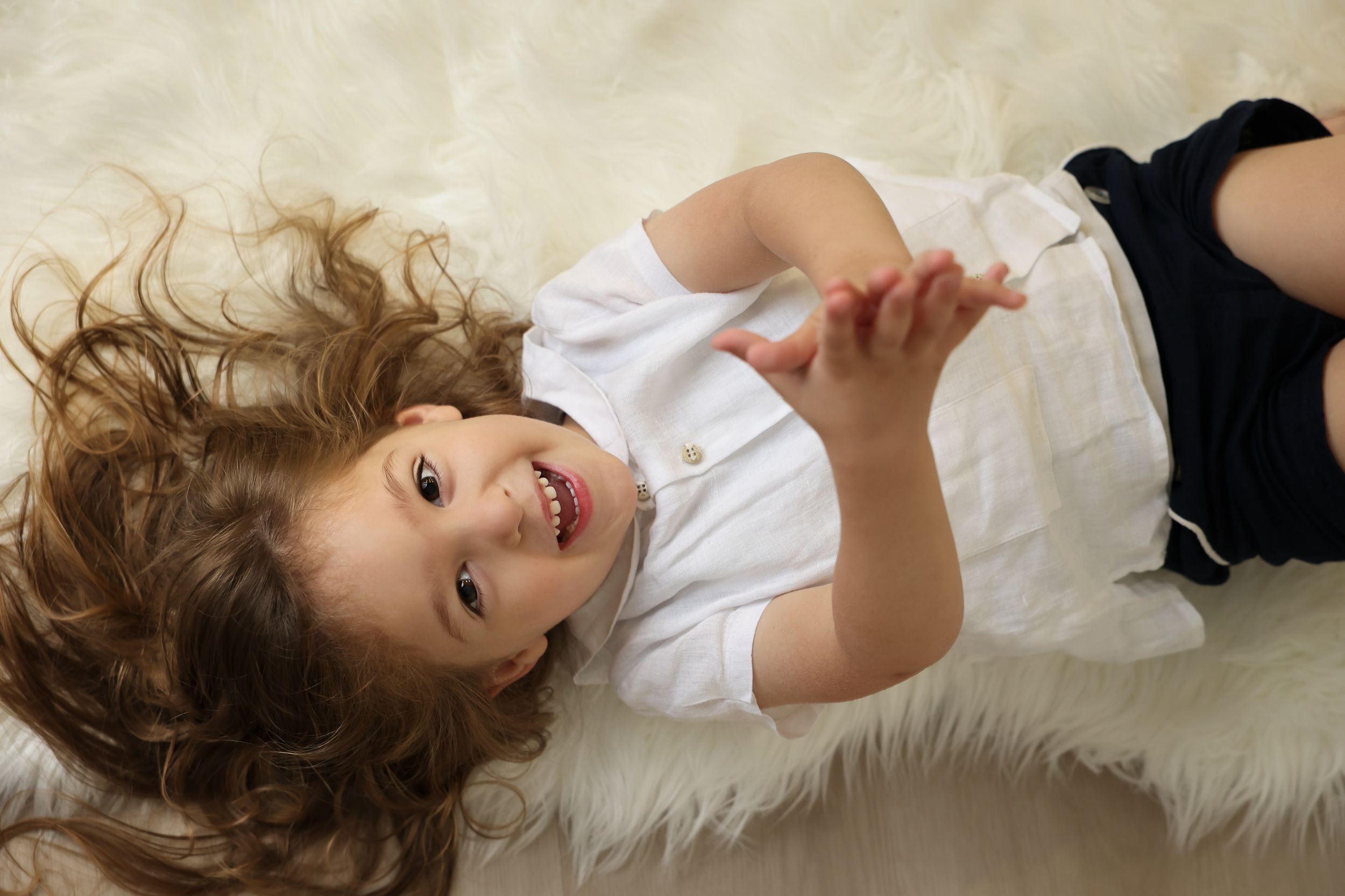 Overhead studio portrait of a happy upsherin boy lying on a white fur rug with his long curly hair fanned out around his head