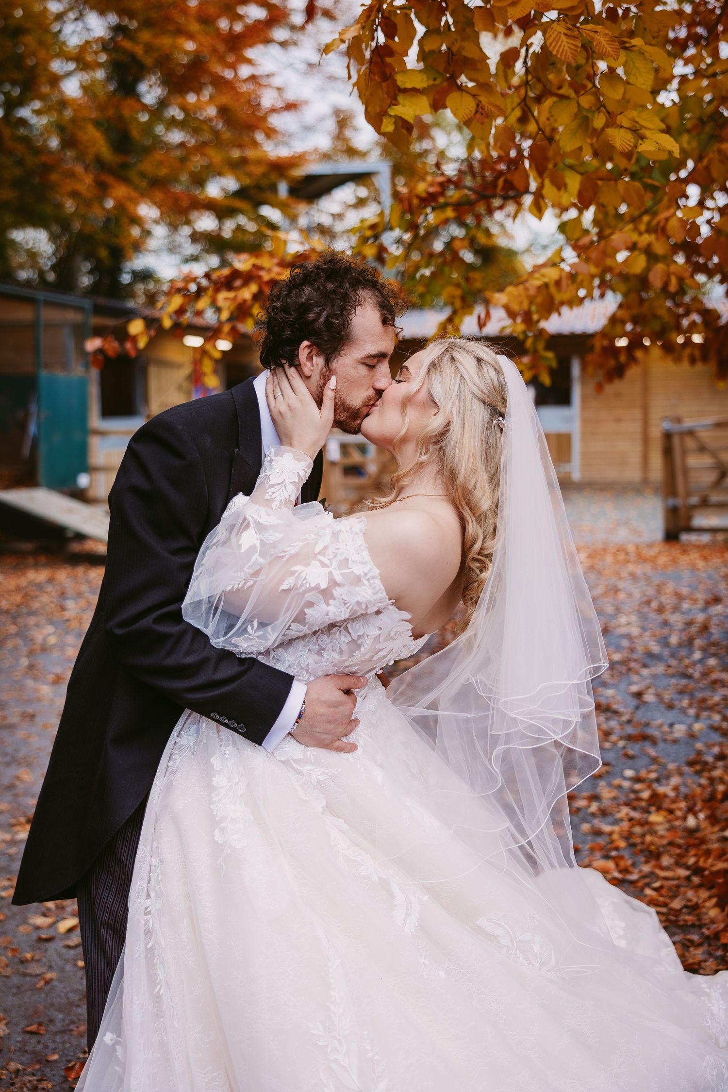 A romantic wedding couple shares intimate kisses under vibrant autumn foliage with golden orange leaves.