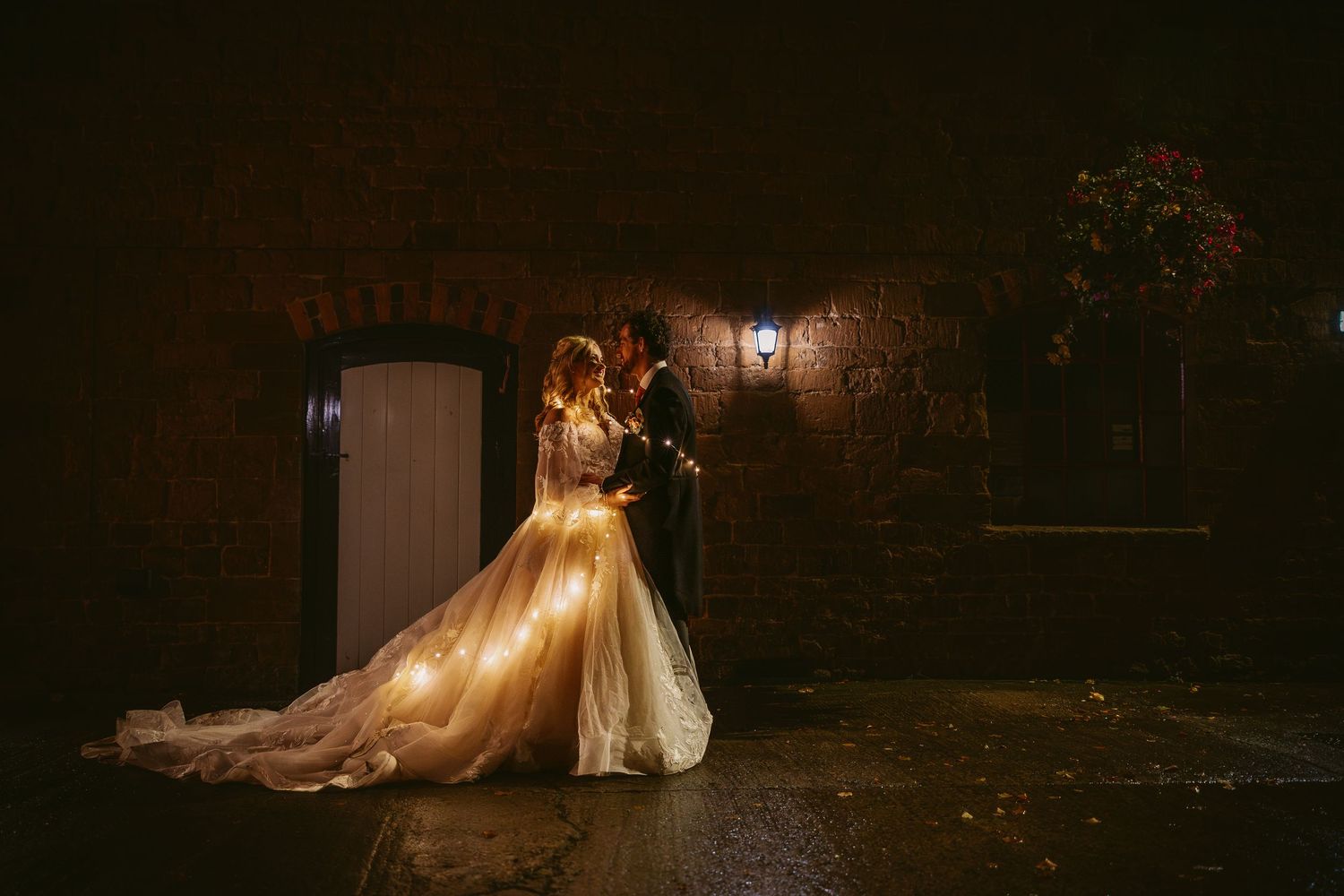 A romantic wedding couple embraces in a glowing evening scene with a flowing champagne-colored gown against dark stone walls.
