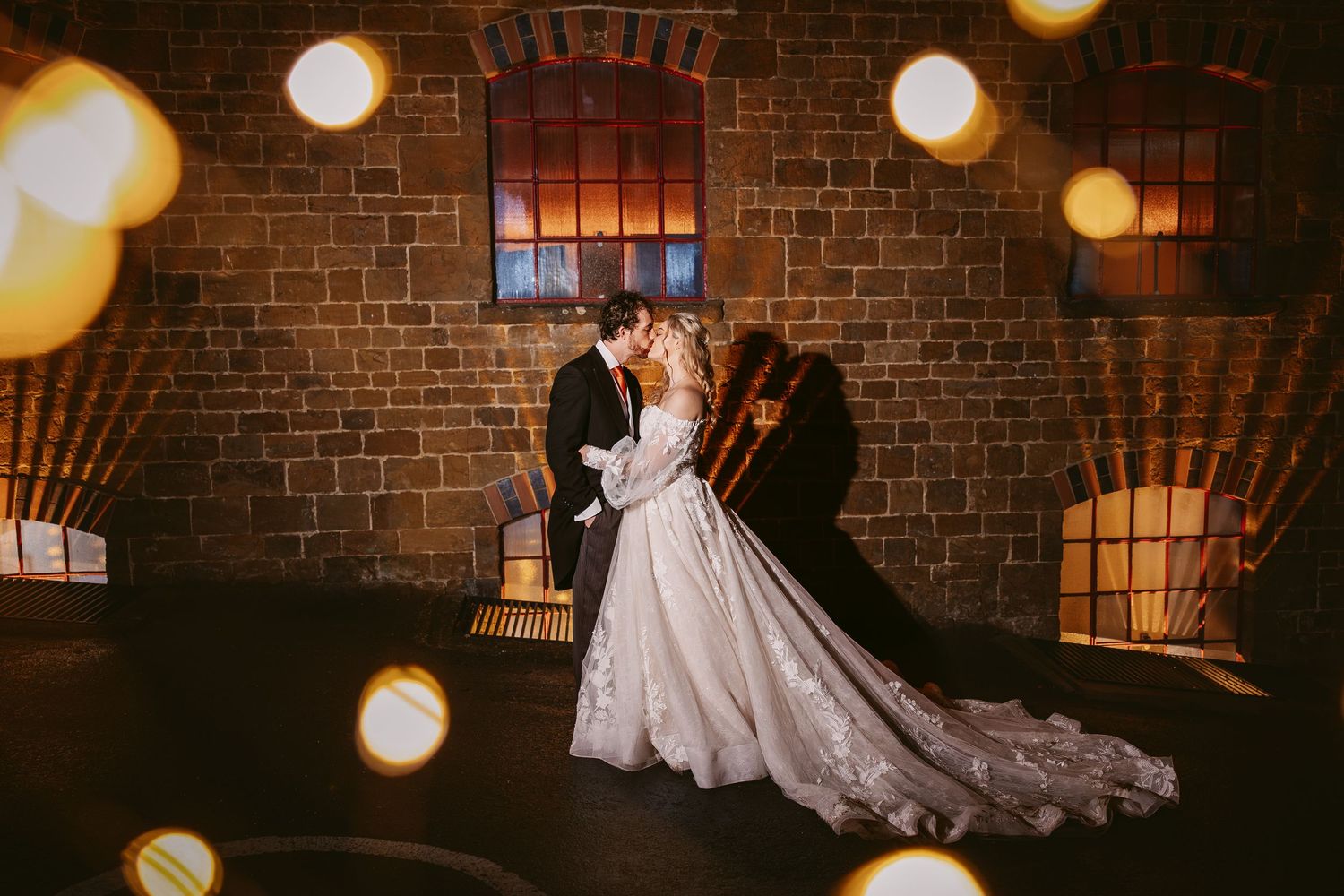 A bride in a flowing white gown stands in a dimly lit brick room with glowing orbs of light creating a romantic ambiance.