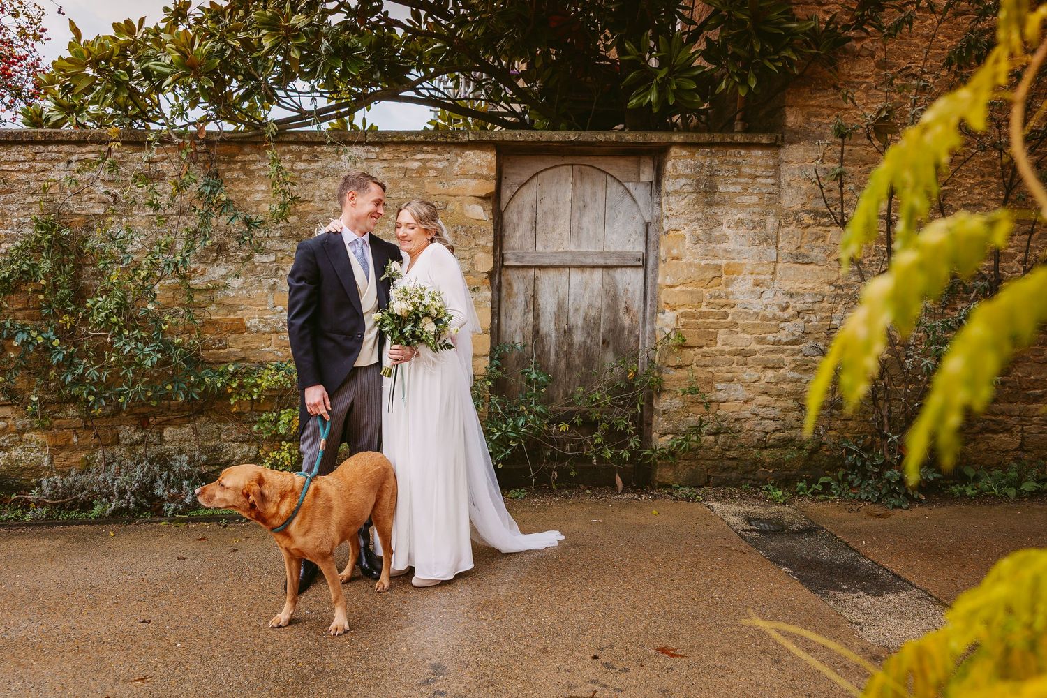 Couple walking along stone wall in garden setting during autumn season with golden foliage overhead.