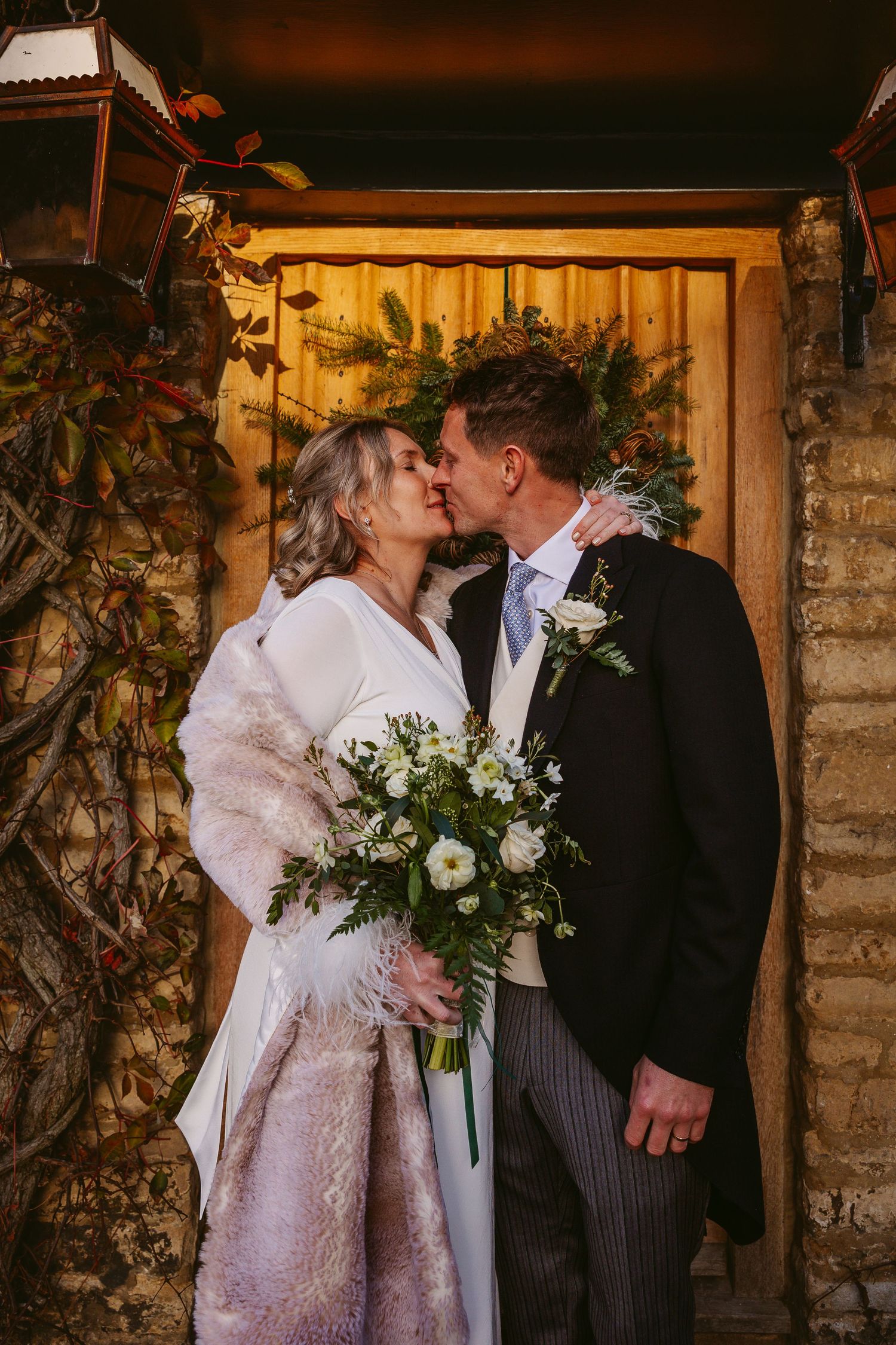 A romantic winter wedding moment captured against a rustic stone wall with white floral bouquet and formal attire.
