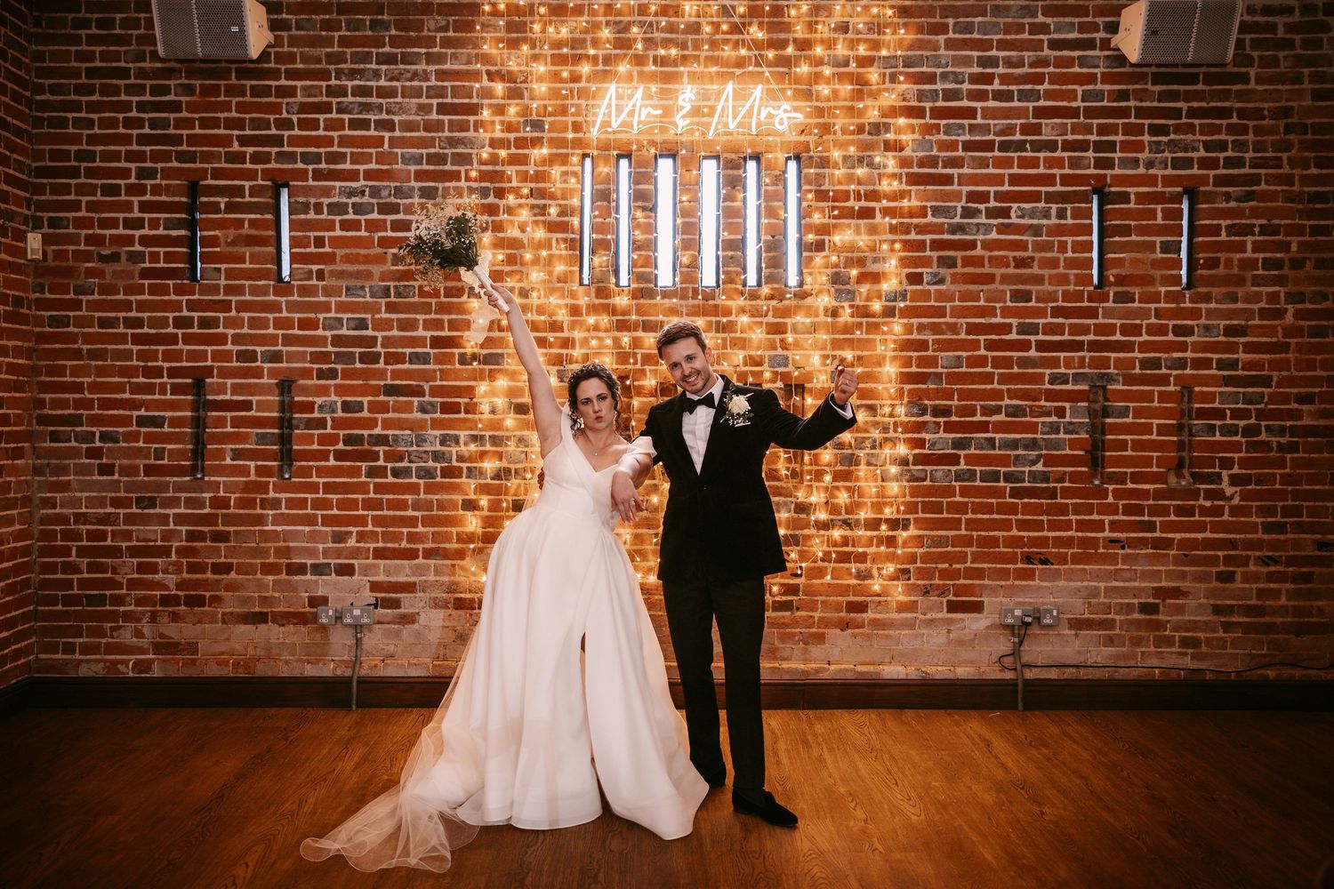 Newlyweds pose against rustic brick wall with elegant lighting in historic venue setting.