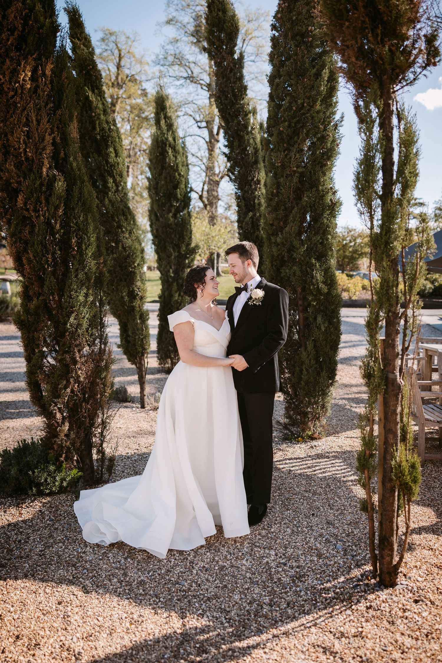 A romantic wedding couple shares intimate moments among tall cypress trees on a gravel path.