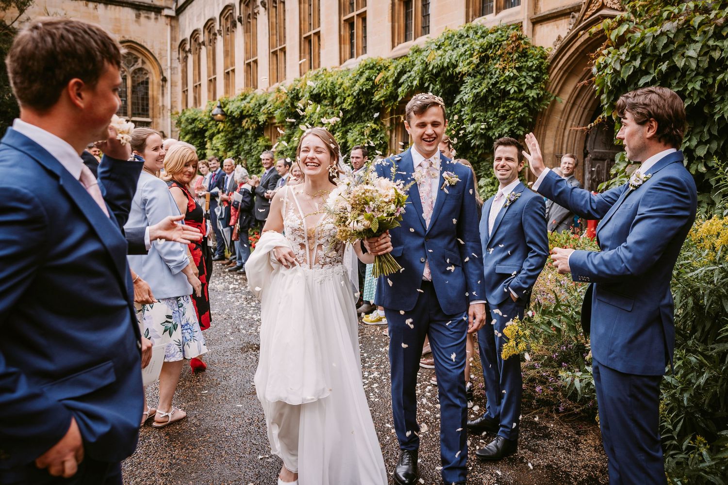 Wedding guests throw confetti over newlyweds outside historic stone building during daytime celebration.