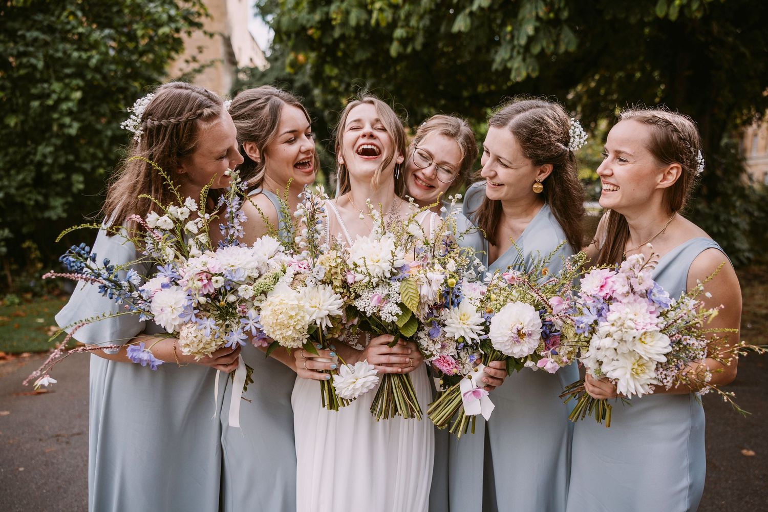 Wedding party members in gray dresses laughing together while holding colorful floral bouquets outdoors.