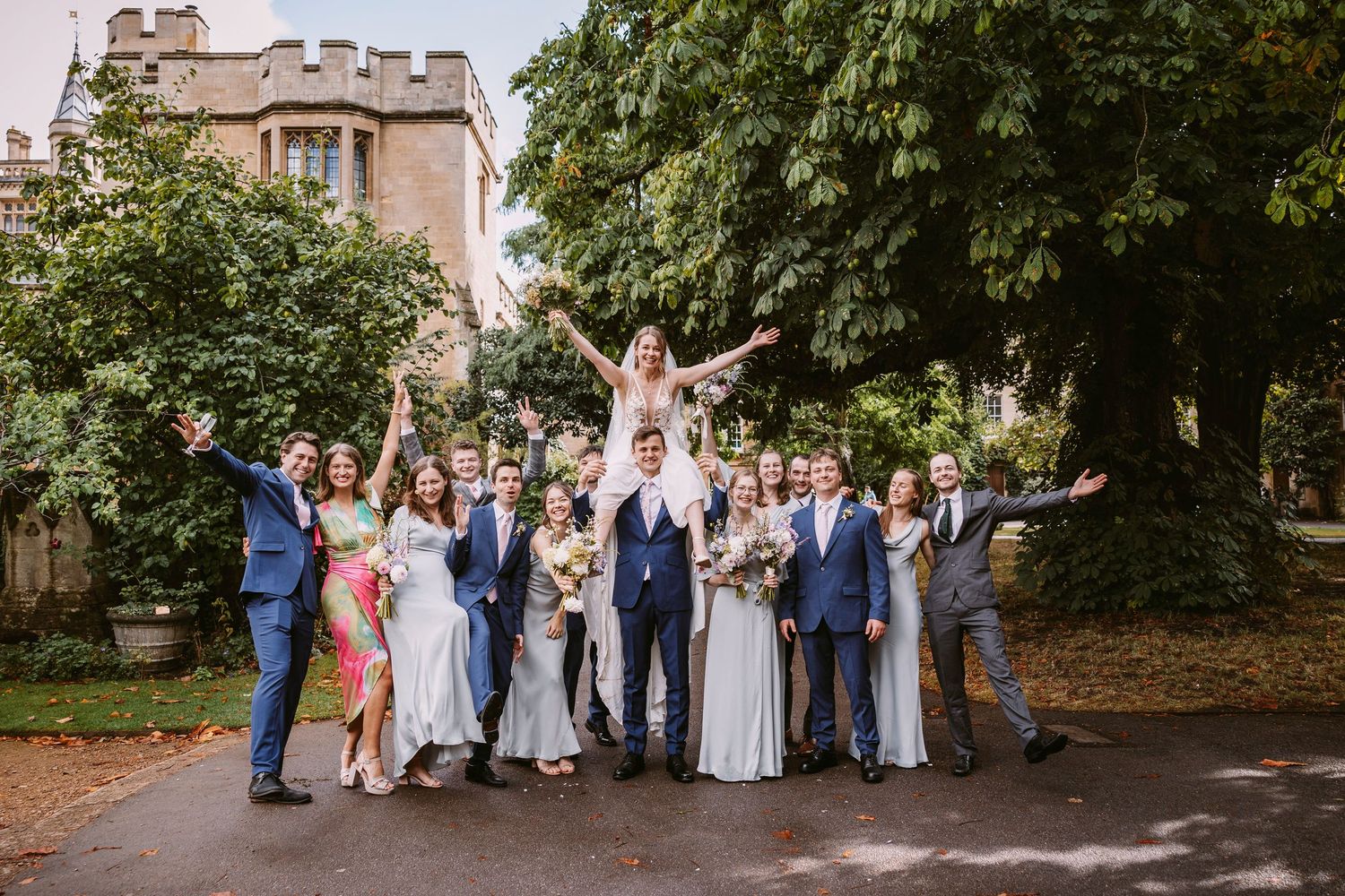 Wedding party walks down a stone path lined with trees at a historic venue during an outdoor celebration.