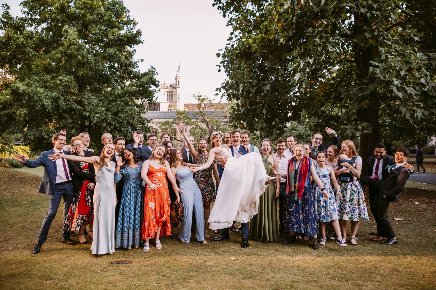 Large group photo of wedding guests gathered outdoors on a sunny day with trees in the background.