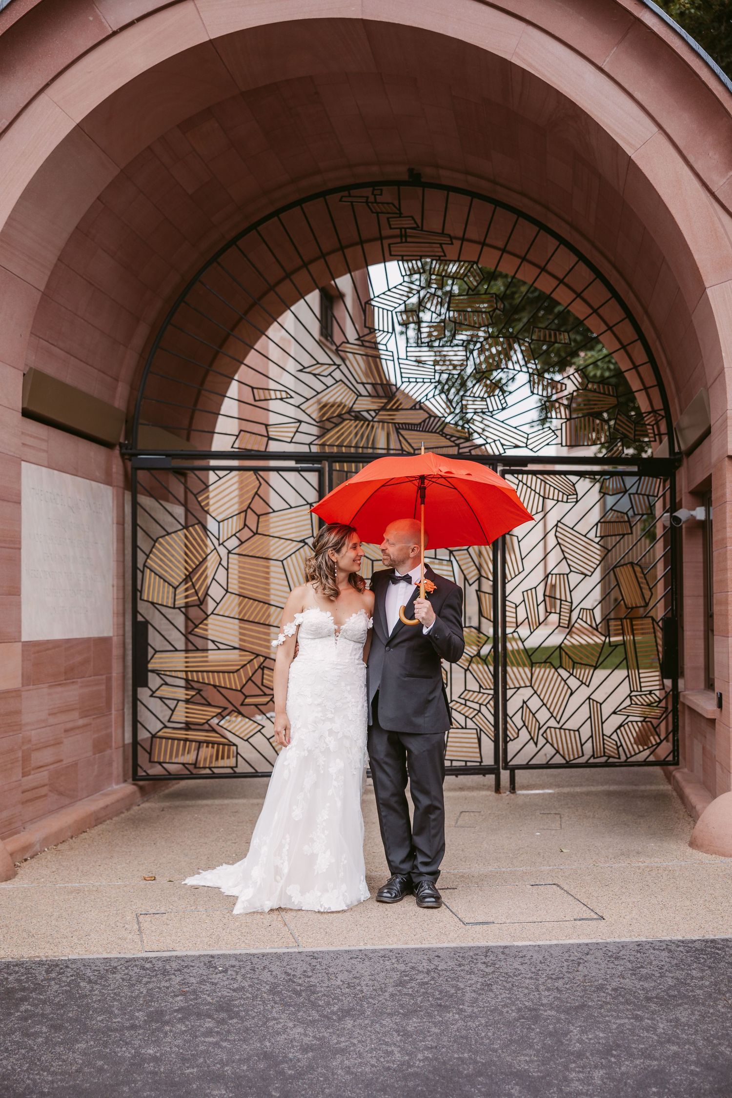 A couple poses under arched walkways with a red umbrella in a series of romantic wedding photos.
