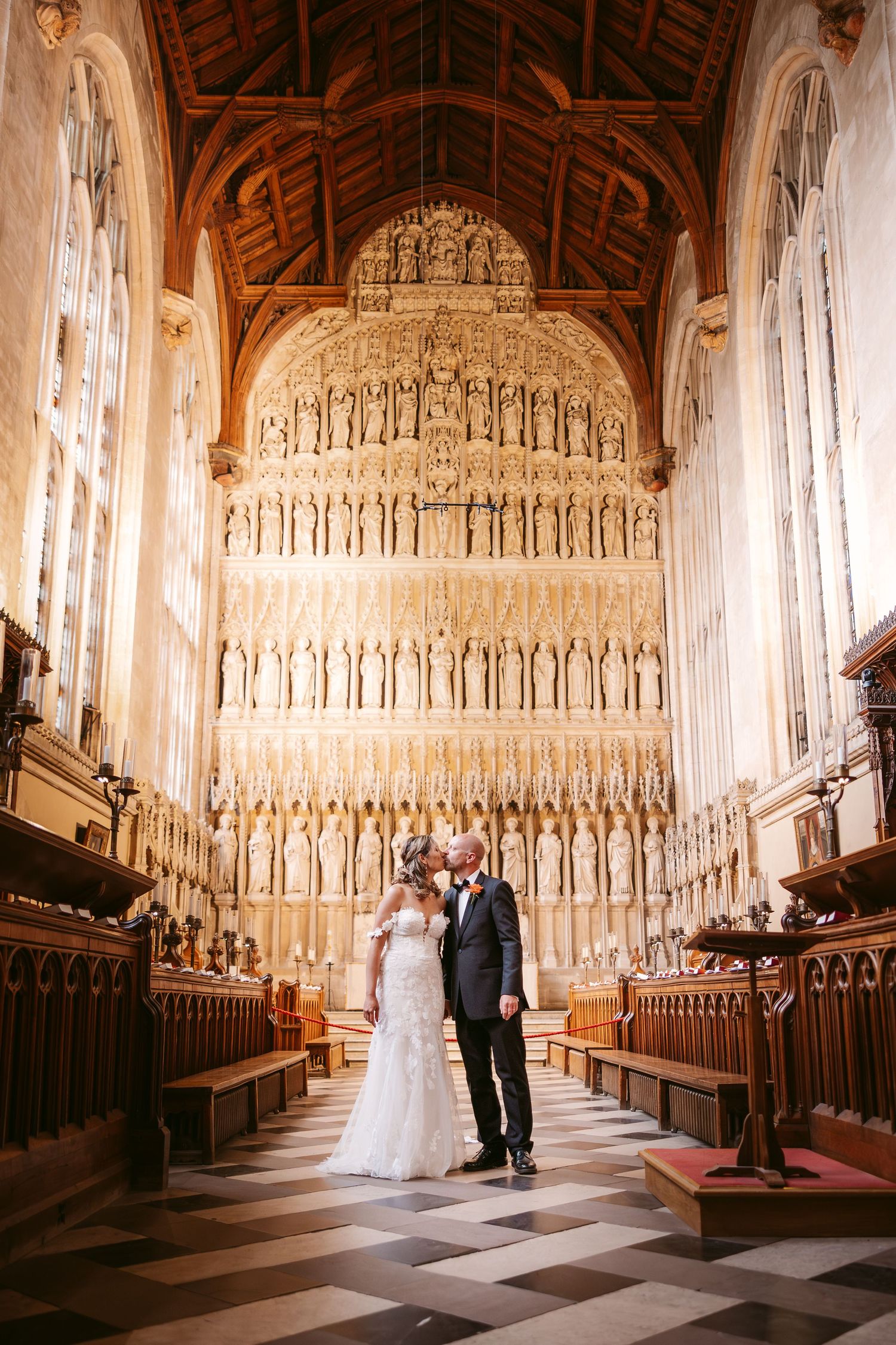 A couple stands in a grand cathedral with ornate wooden beams and a stunning tiered stone reredos wall behind them.