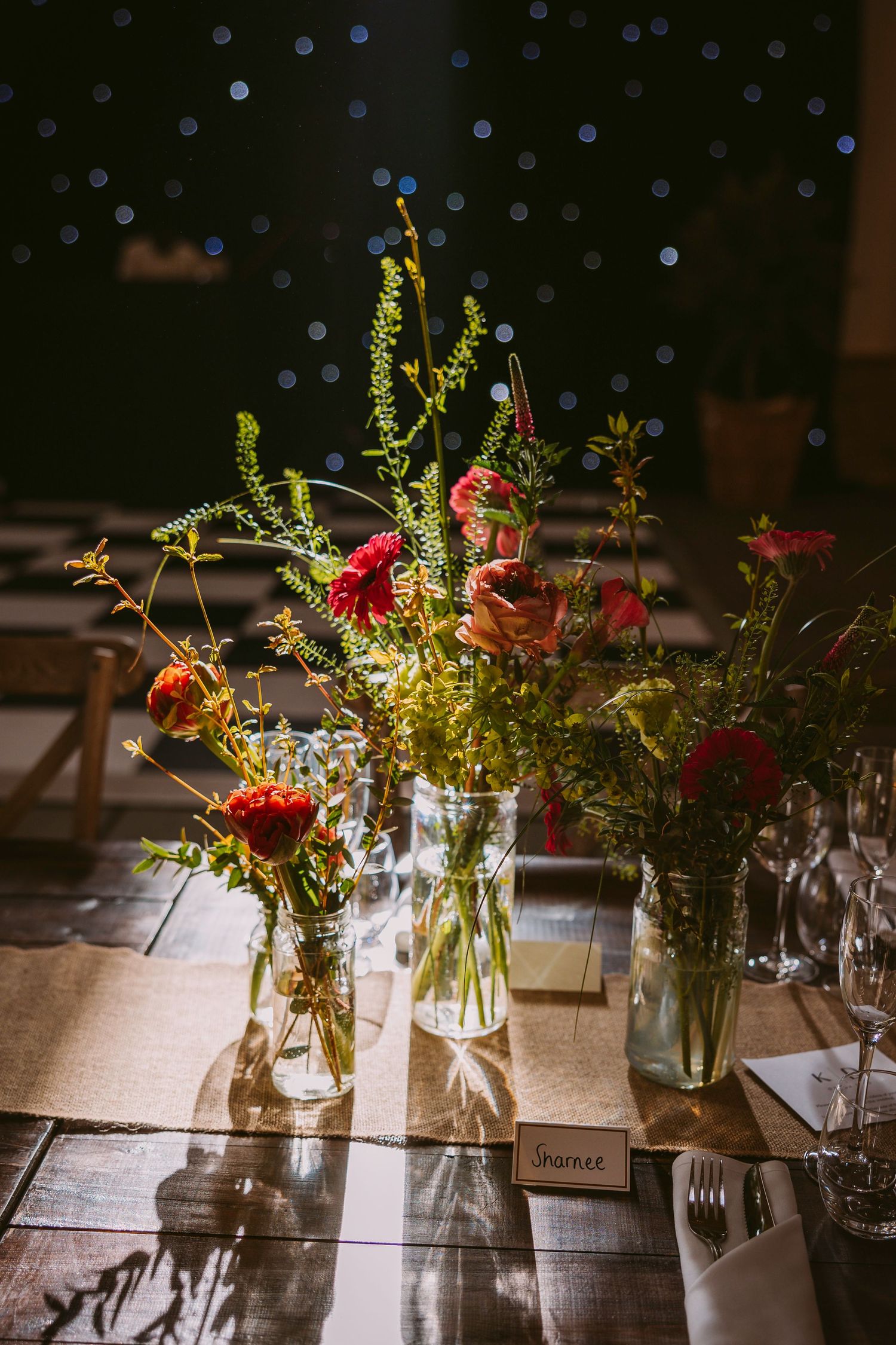 Simple glass jar centerpieces with wildflowers and greenery arranged on rustic wooden tables at a wedding reception.
