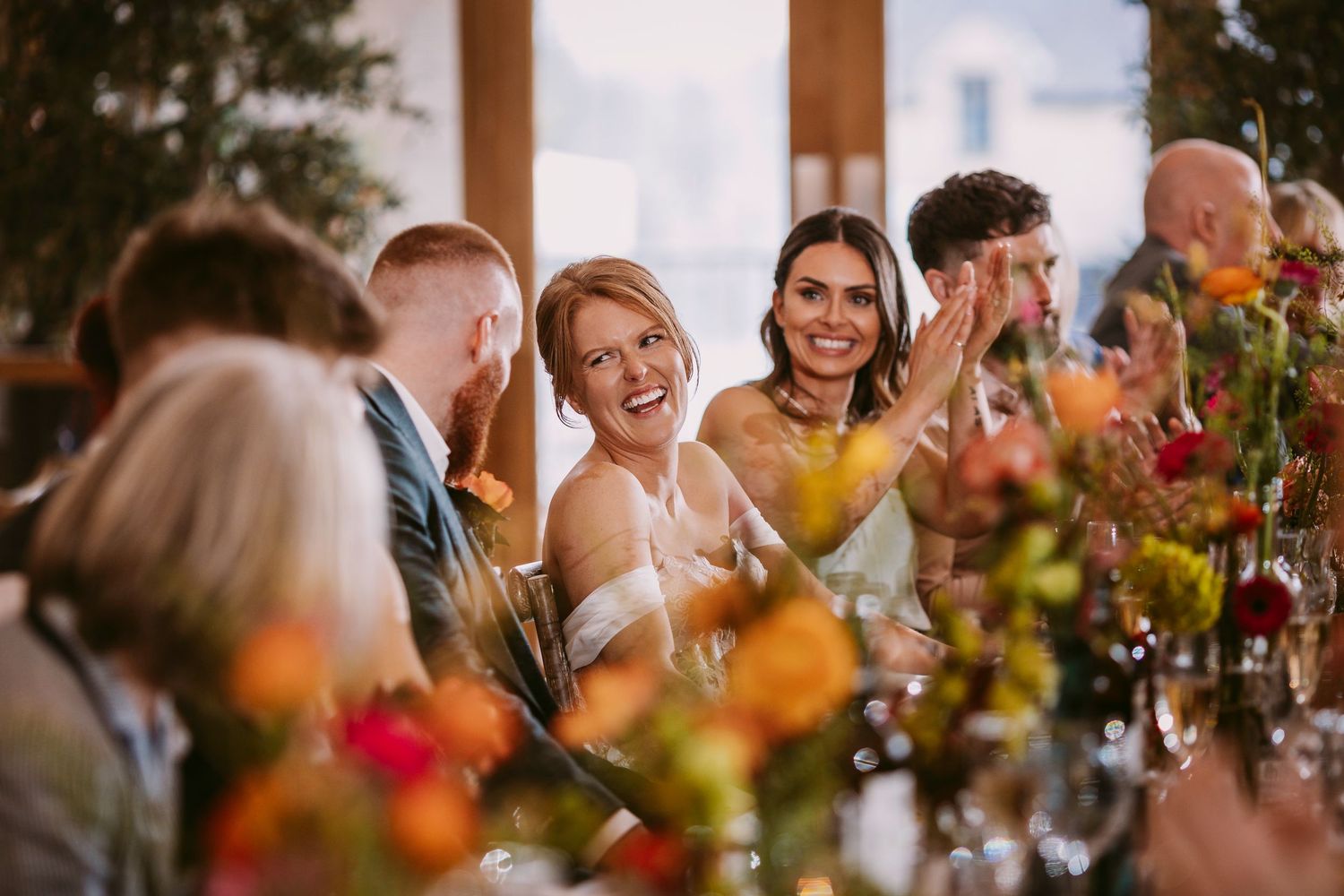Wedding guests laugh and smile during a reception dinner surrounded by colorful floral centerpieces.