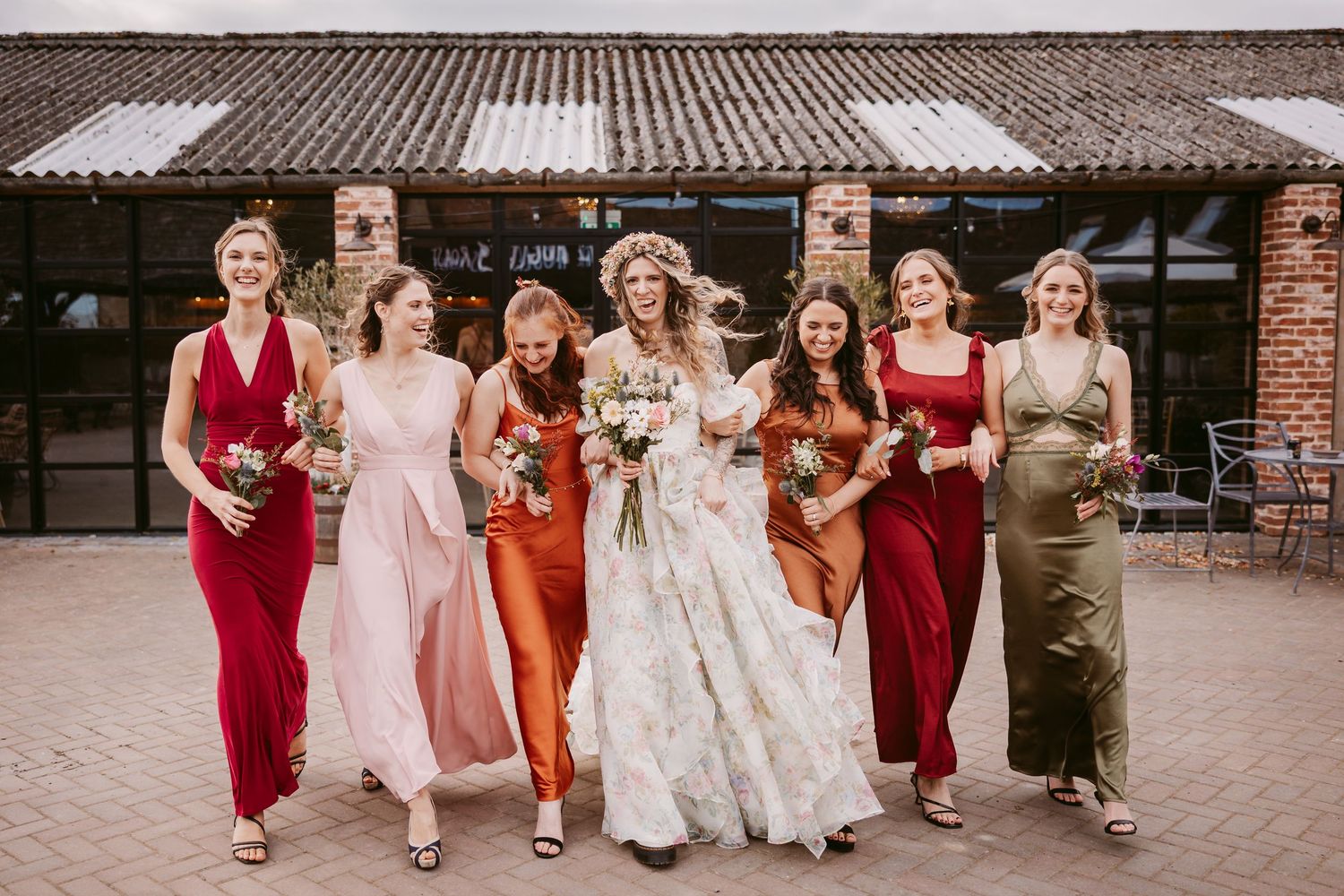 Wedding party wearing burgundy and rust colored dresses walk together on brick pathway at outdoor venue.