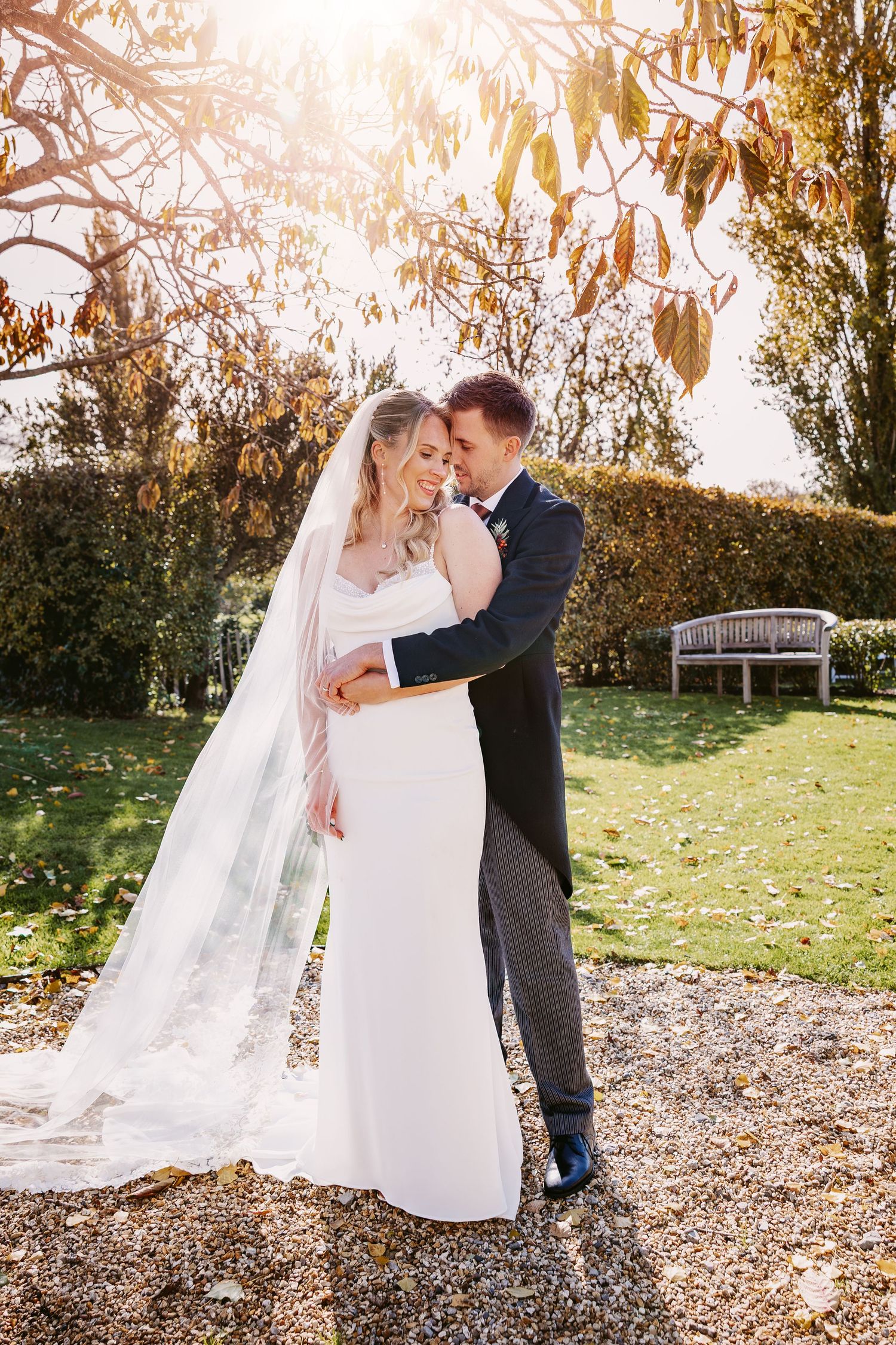 A romantic wedding couple embraces in a sunlit autumn garden with fallen leaves and a flowing white veil at Merriscourt in the Cotswolds