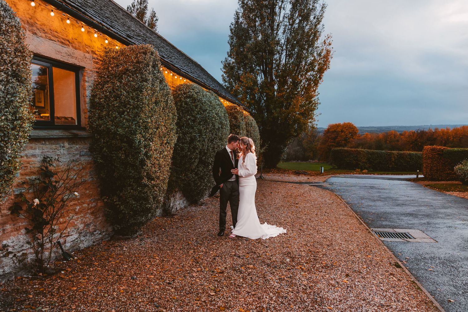 Bride and groom embrace in autumn wedding photo shoot along a tree-lined path by the water at sunset.