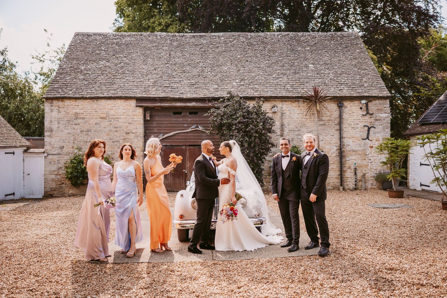 Wedding party poses for photos outside rustic stone barn venue at sunset.